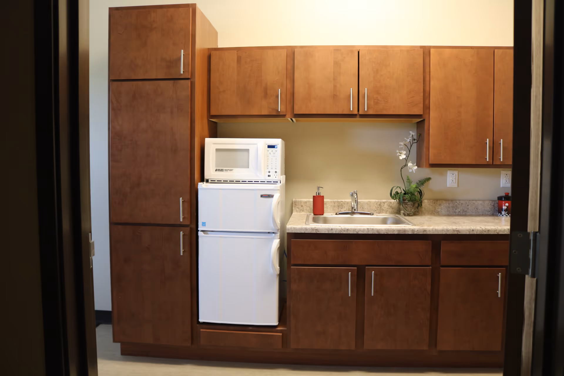 A small kitchen area with wooden cabinets, a white microwave on top of a white mini refrigerator, a countertop with a sink, a red soap dispenser, and a small potted plant with white flowers.