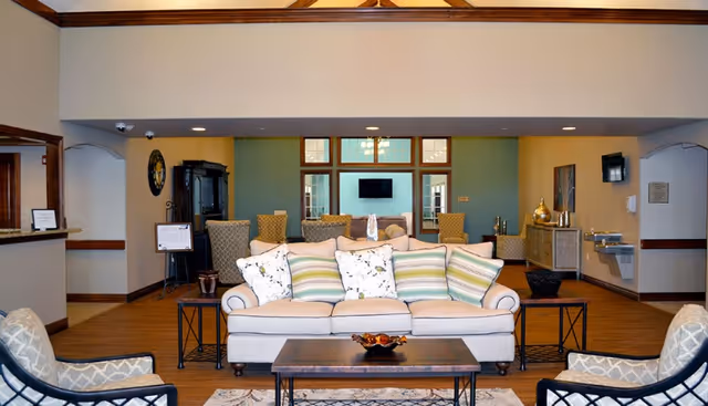 A spacious and well-lit living room area with a beige sofa adorned with striped and floral cushions, two patterned armchairs, a wooden coffee table with a decorative bowl, and additional seating in the background. The room features wooden flooring, a wall-mounted TV, decorative items on side tables, and a water fountain on the right wall.