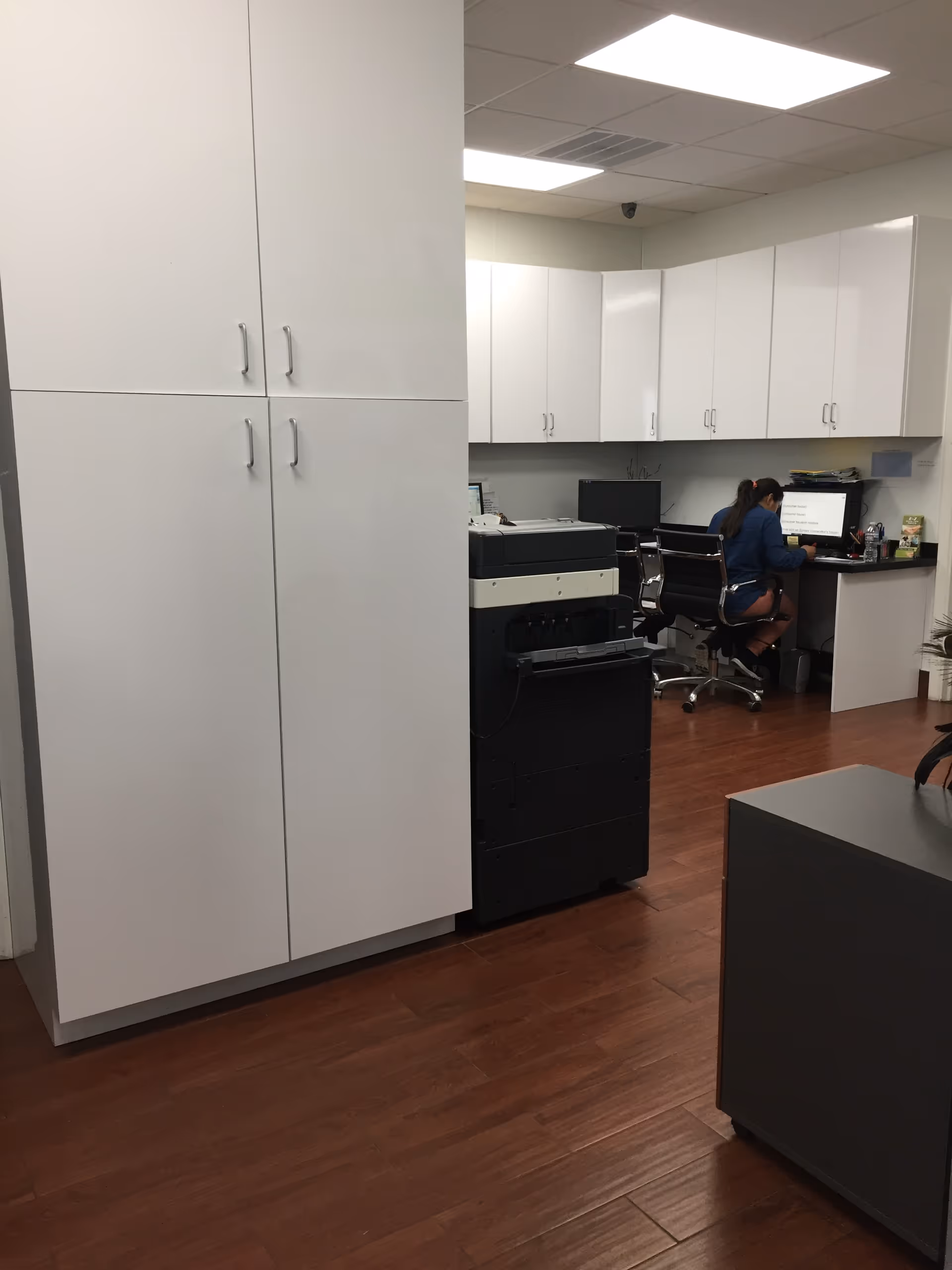 White cabinets surround a small office area with a copier and two people at desks