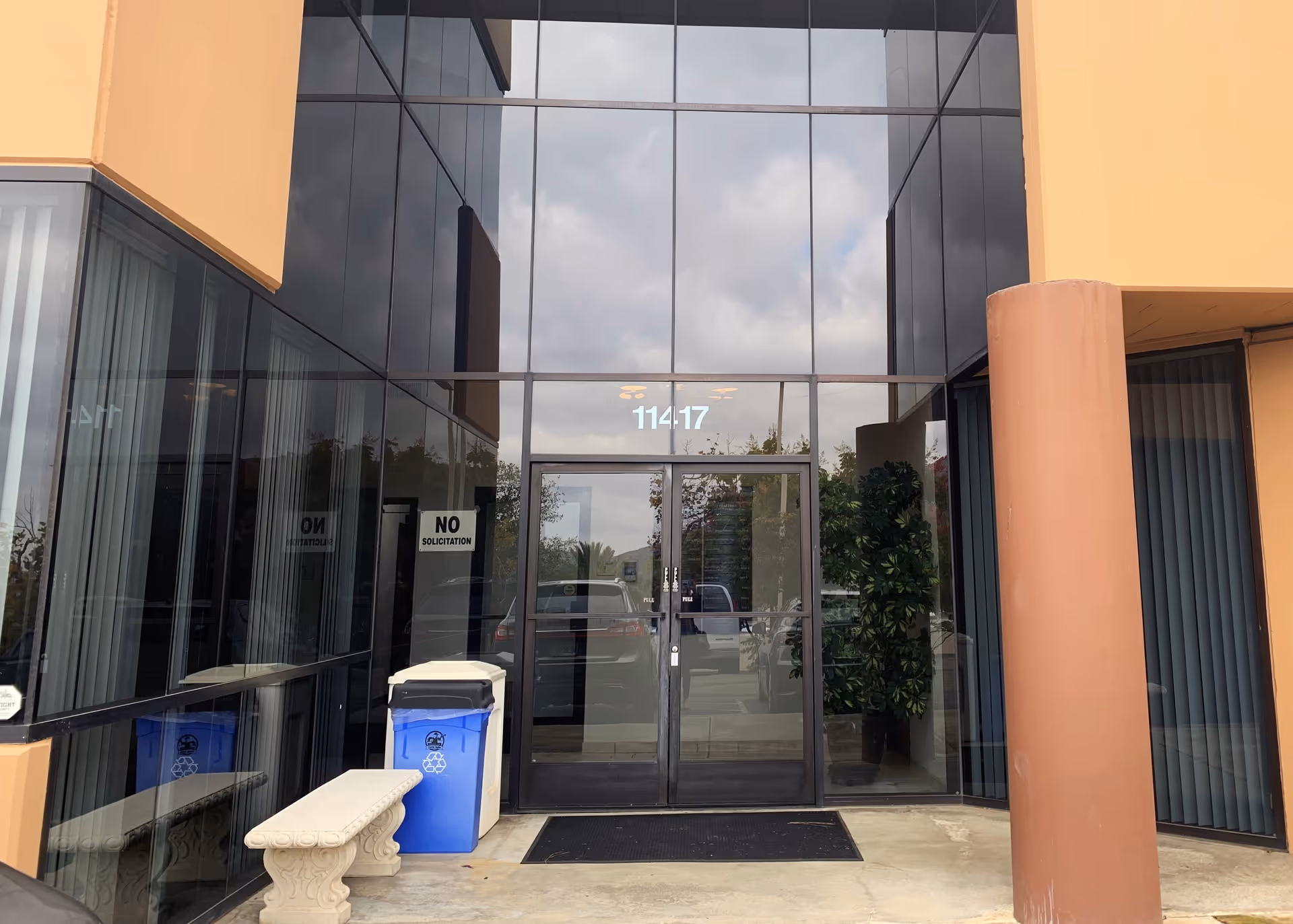 Glass-front entrance with double doors, bins, and a bench outside the building.
