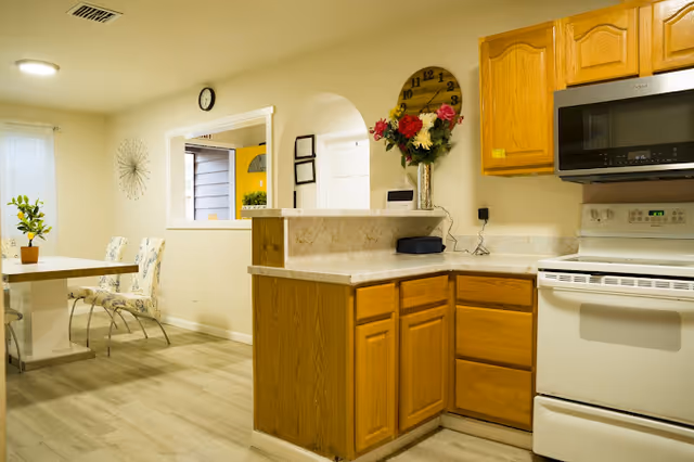 A compact kitchen and dining area with wooden cabinets, stove, and flower centerpiece.