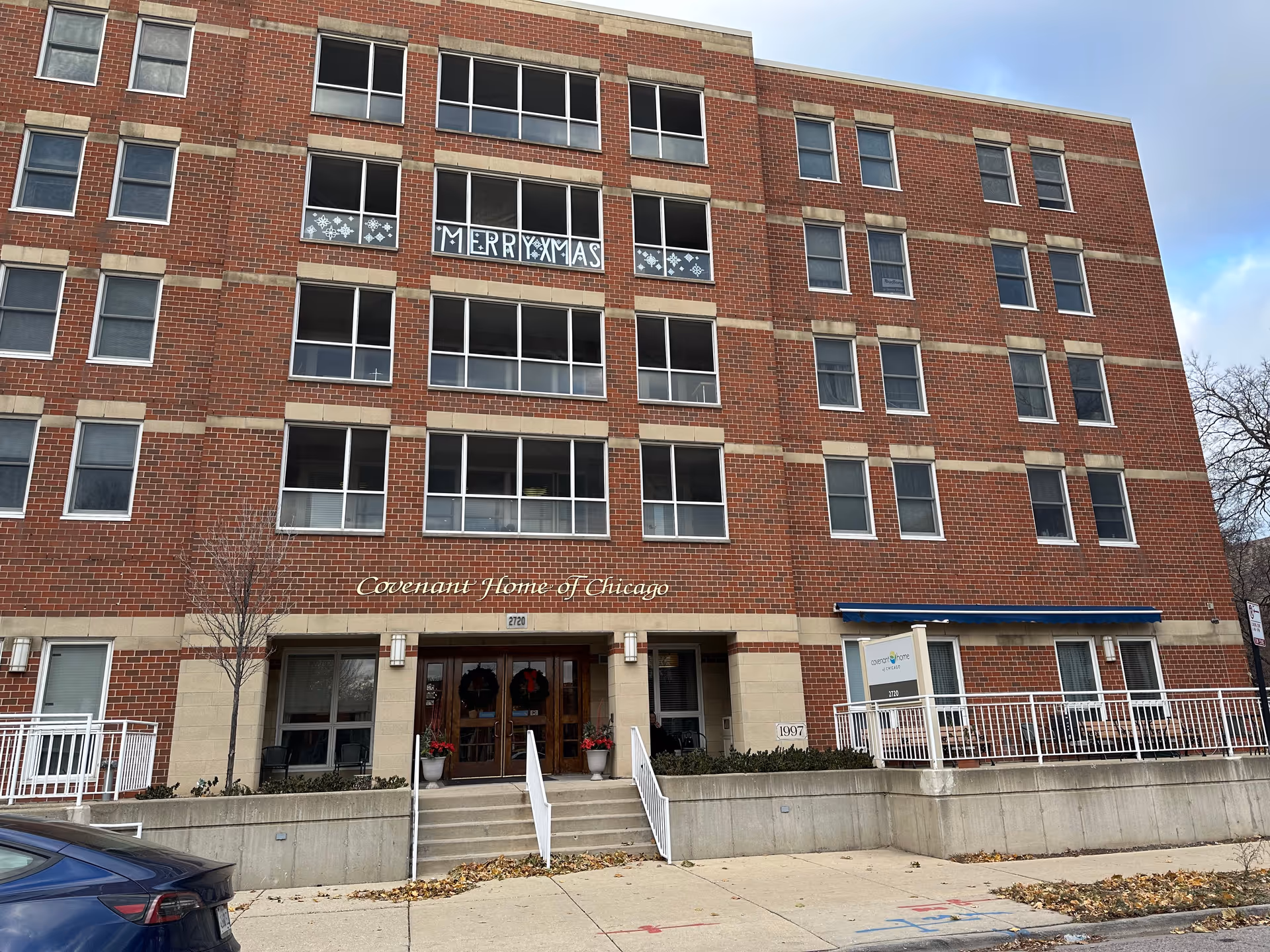 Exterior view of a multi-story brick building with the sign 'Covenant Home of Chicago' above the entrance. The entrance has double wooden doors decorated with wreaths and potted plants on either side. Several windows are visible, with one window displaying a festive 'MERRY XMAS' sign and snowflake decorations. A blue car is parked in front of the building, and a sidewalk runs along the front.