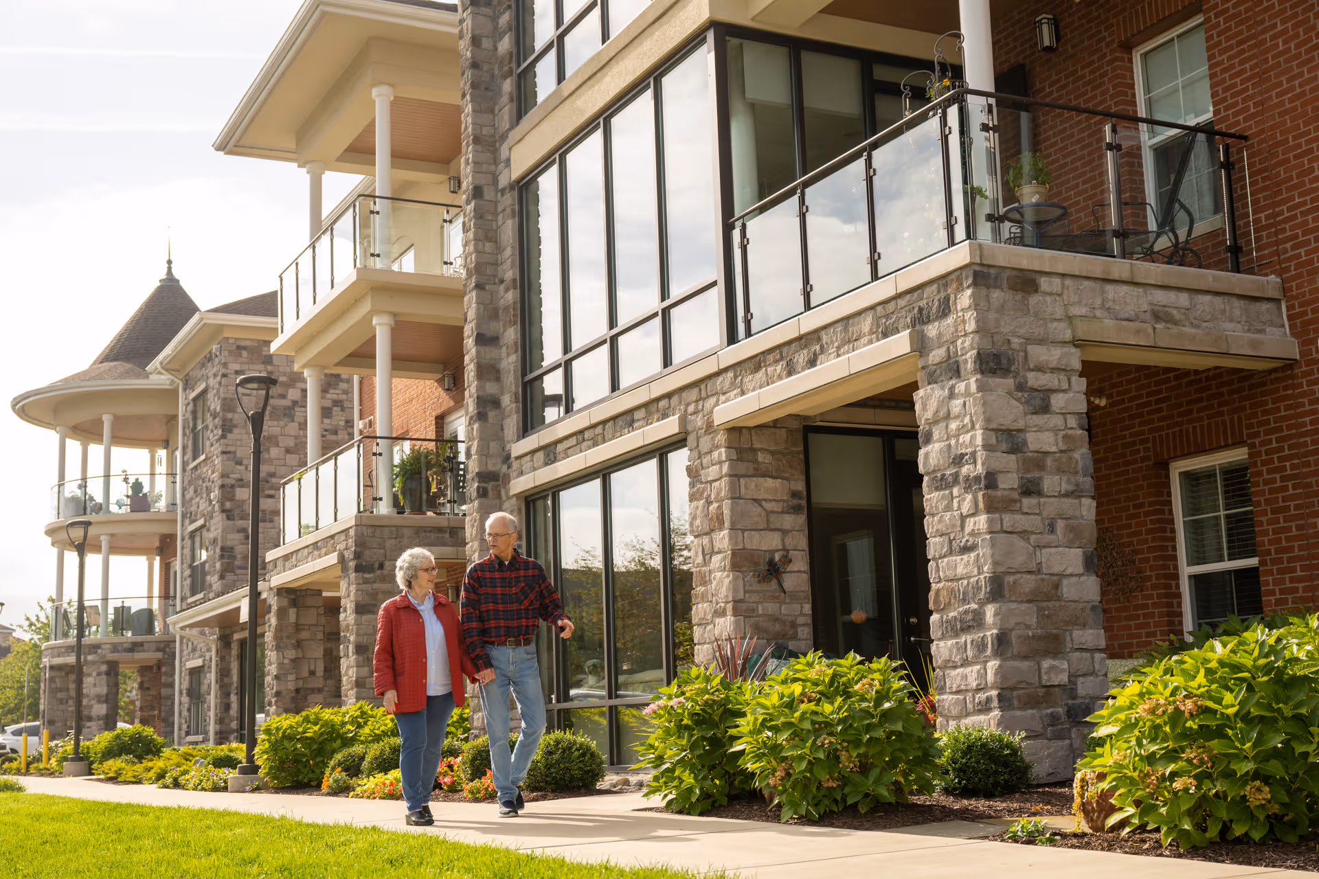 An elderly couple walking on a sidewalk outside a modern senior living facility with stone and brick exterior, balconies with glass railings, and well-maintained landscaping.