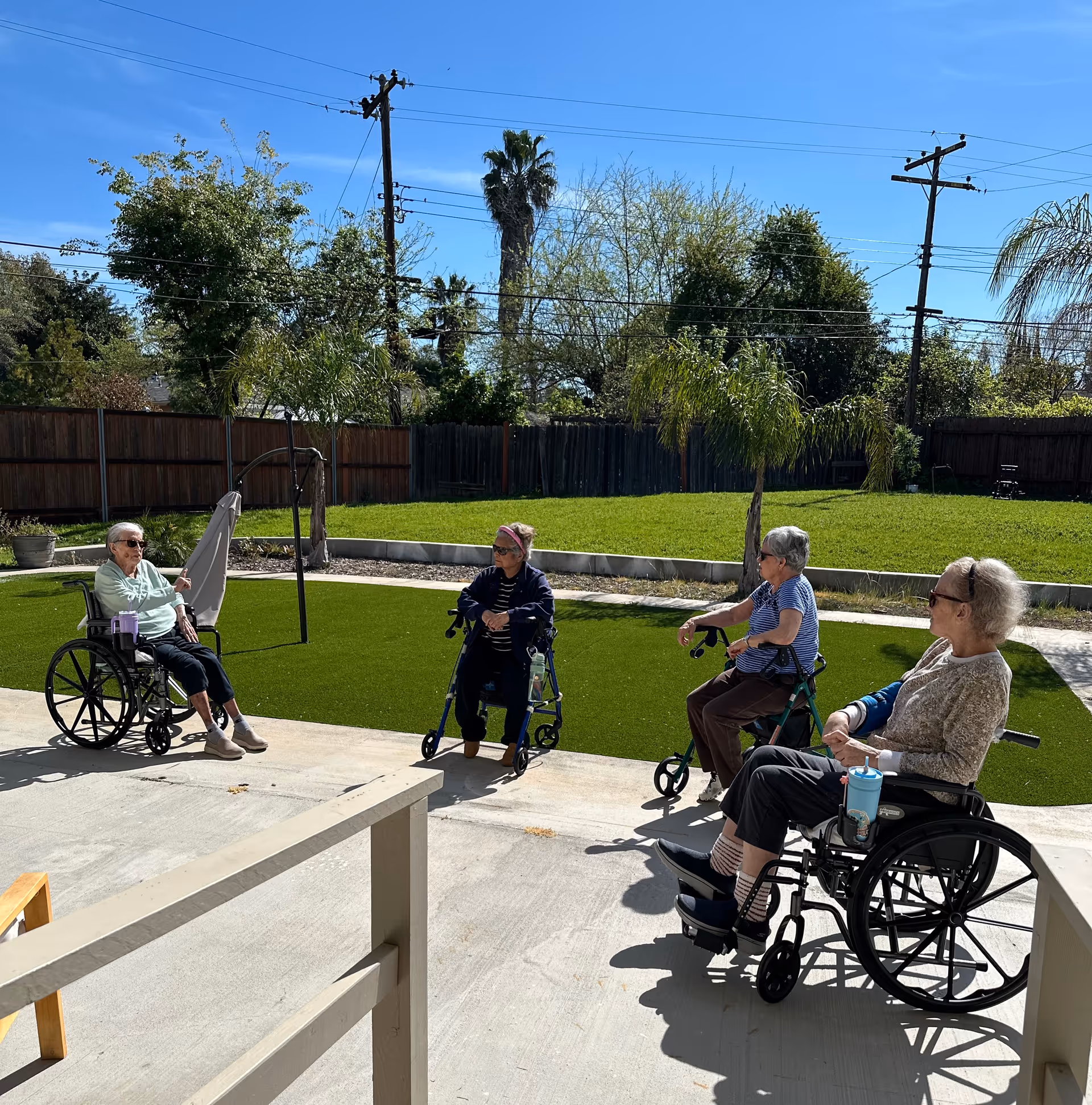 Four elderly residents in wheelchairs and a walker sit in a sunny outdoor courtyard.