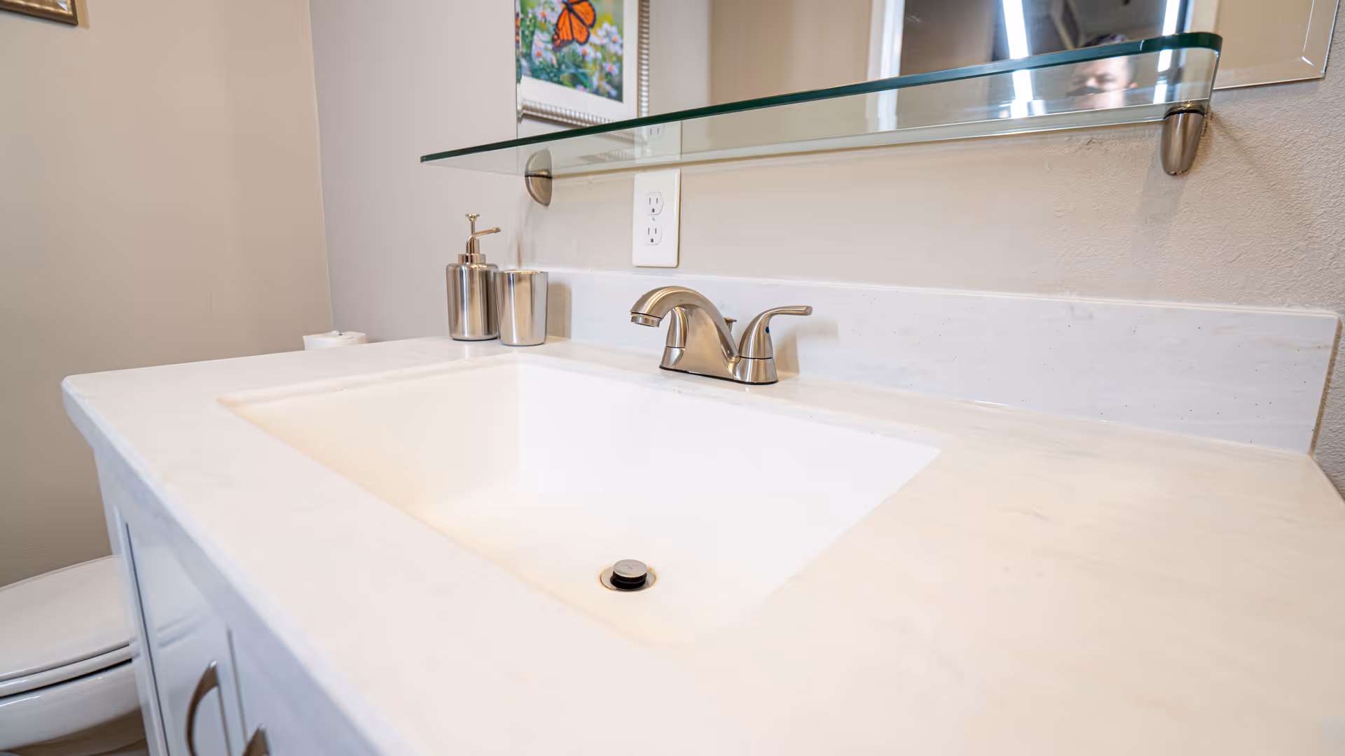 A clean bathroom sink with a silver faucet, soap dispenser, and cup on a white countertop. A toilet is partially visible to the left, and a mirror with a glass shelf is mounted on the wall above the sink. A colorful butterfly picture is reflected in the mirror.