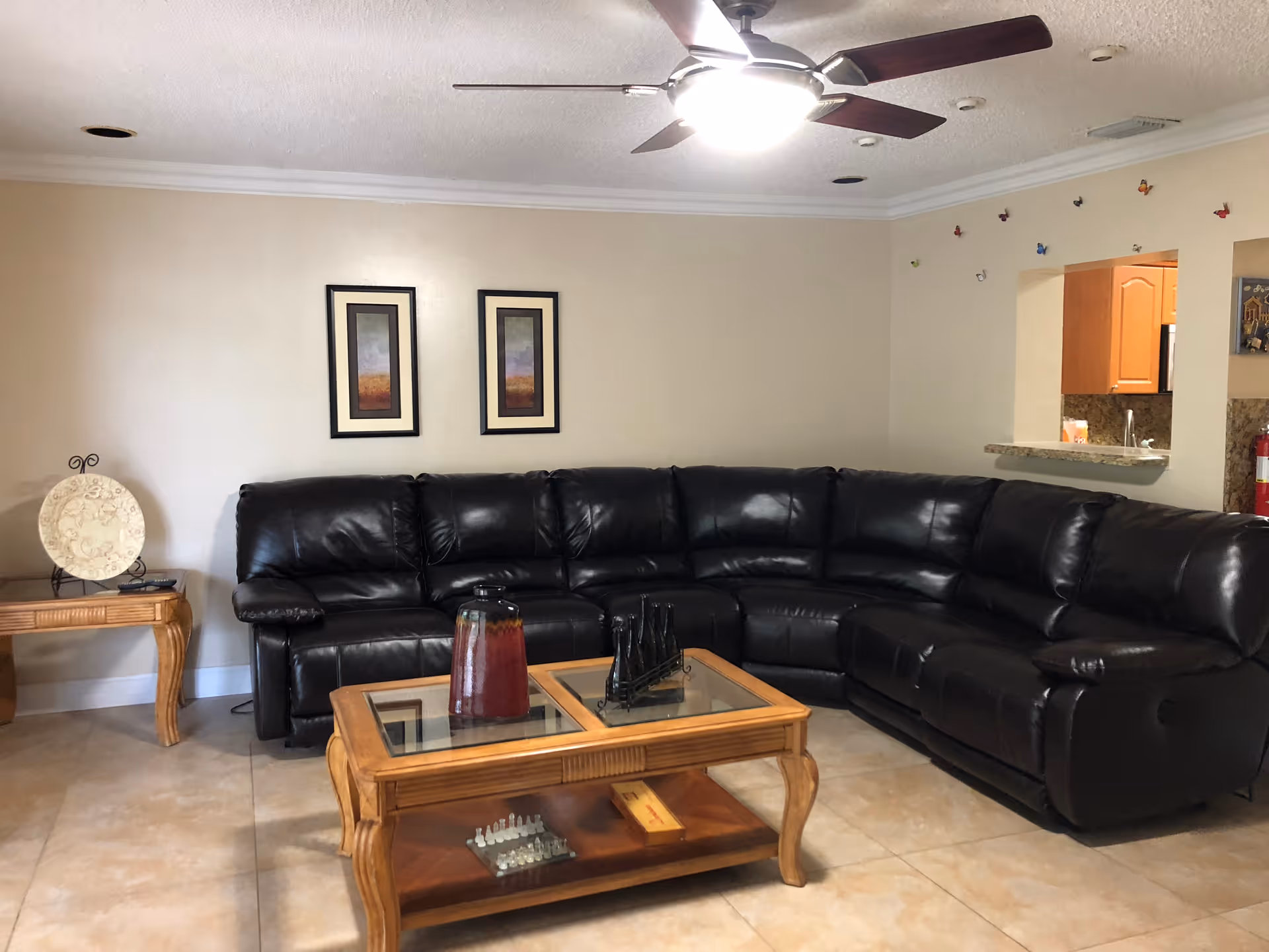 A living room with a large black leather sectional sofa, a wooden coffee table with glass inserts and decorative items on top, a side table with a decorative plate, two framed pictures on the wall, a ceiling fan with light, and a pass-through window to a kitchen area.