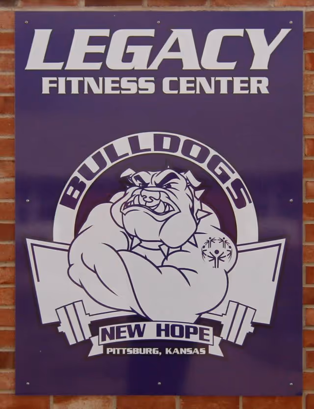 Purple sign reading Legacy Fitness Center with bulldog logo and New Hope on brick wall.