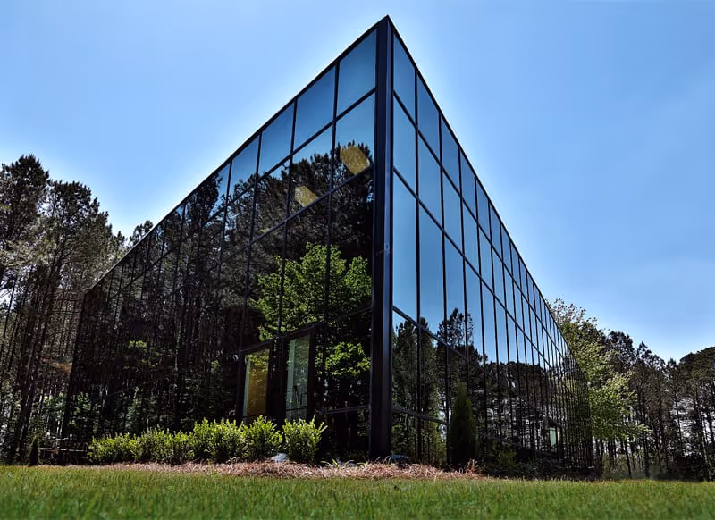 Glass-walled modern building exterior surrounded by trees and a green lawn.