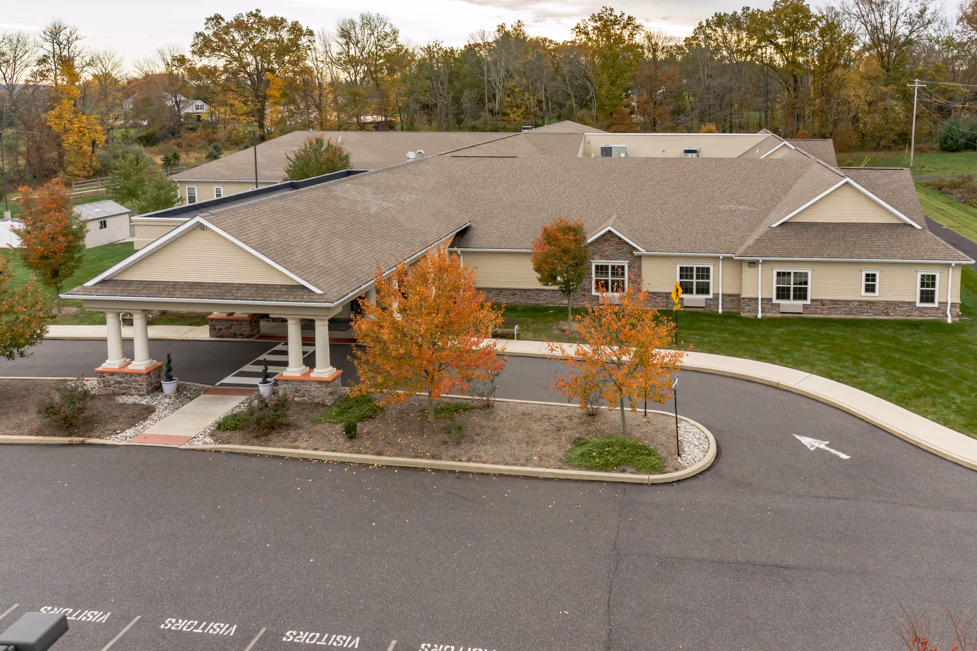 Exterior view of a single-story senior living facility building with a covered entrance, surrounded by autumn trees with orange and yellow leaves, a curved driveway, and a parking area marked for visitors.