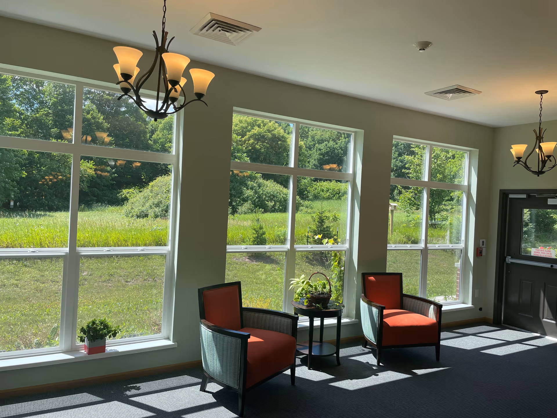 A bright sitting area inside Shawano Place Senior Living with large windows showing a green outdoor landscape. The room has two orange cushioned chairs with dark wooden frames, a small round table with a plant basket between them, and two hanging light fixtures with multiple lamps. A door is visible on the right side.