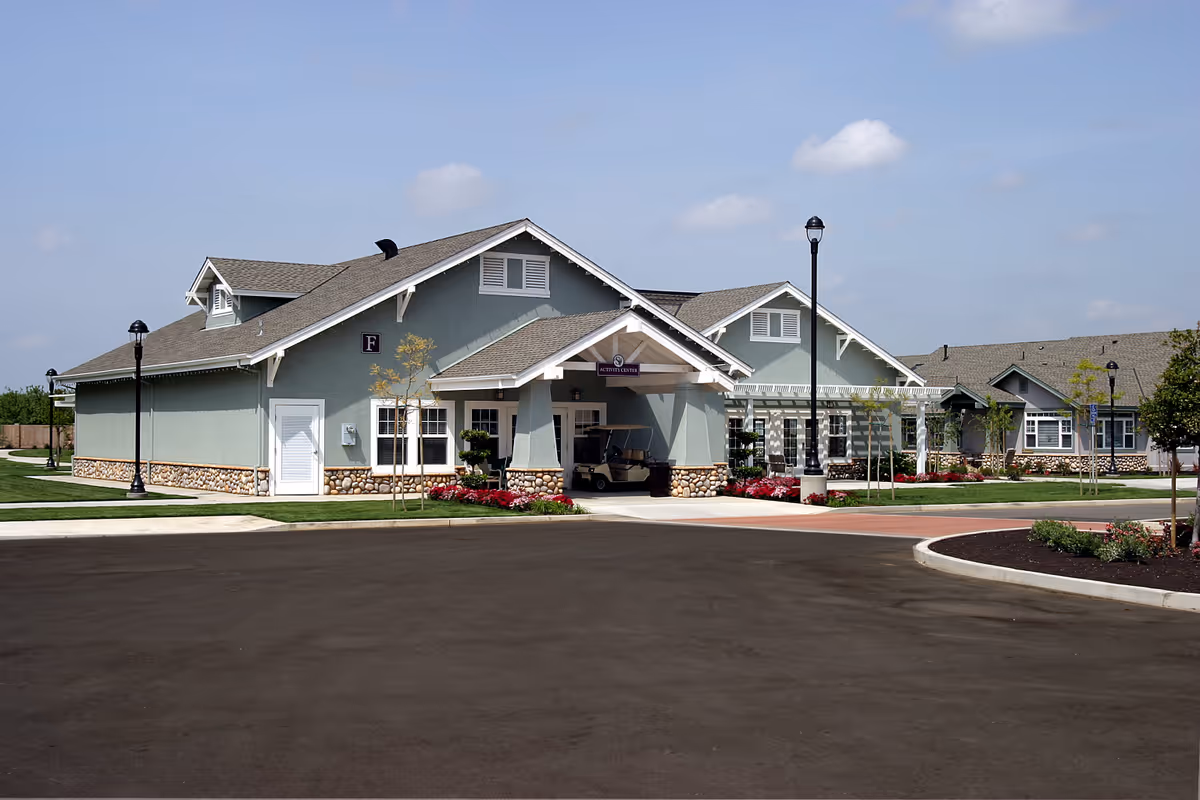 Single-story pale green senior living facility building with a covered main entrance, stone trim, lamp posts, and landscaped grounds.