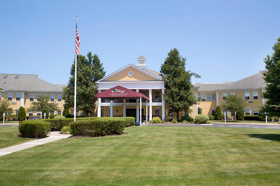Front exterior view of The Village at Buckland Court, a two-story yellow building with white columns and a maroon canopy over the entrance. There is a well-maintained lawn, bushes, trees, and an American flag on a flagpole in front of the building under a clear blue sky.