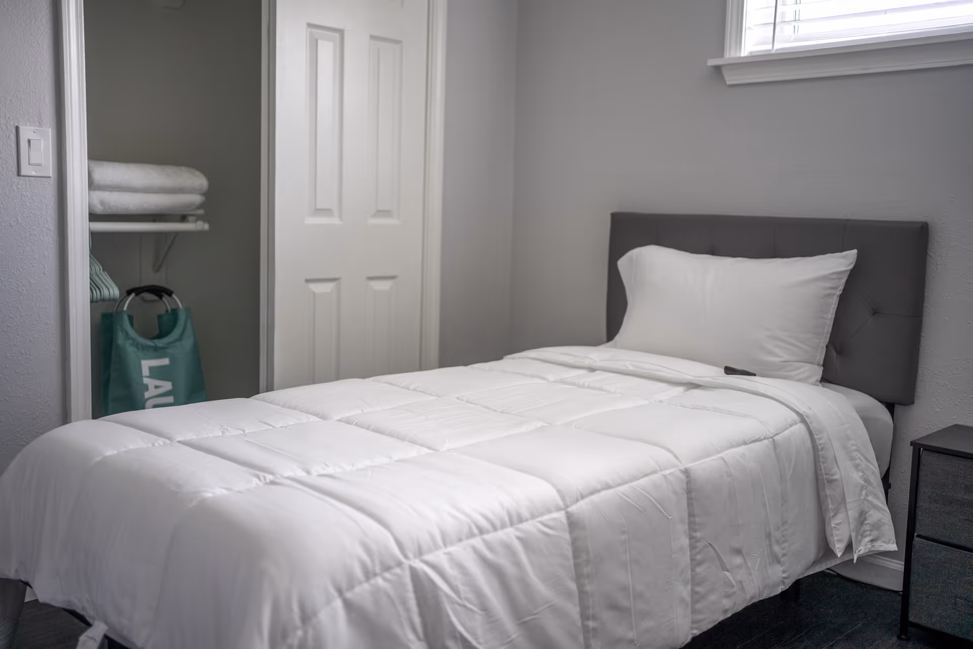 Single bed with white comforter in a small gray bedroom, open closet with towels.