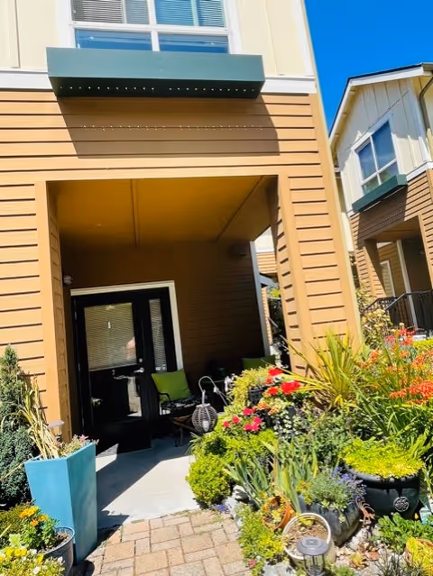 Entrance to a residential building with a covered porch area featuring outdoor seating. The building has tan siding with white trim and a green awning above the door. In front of the entrance, there is a garden with various colorful flowers and green plants in pots and planted beds. The sky is clear and blue.