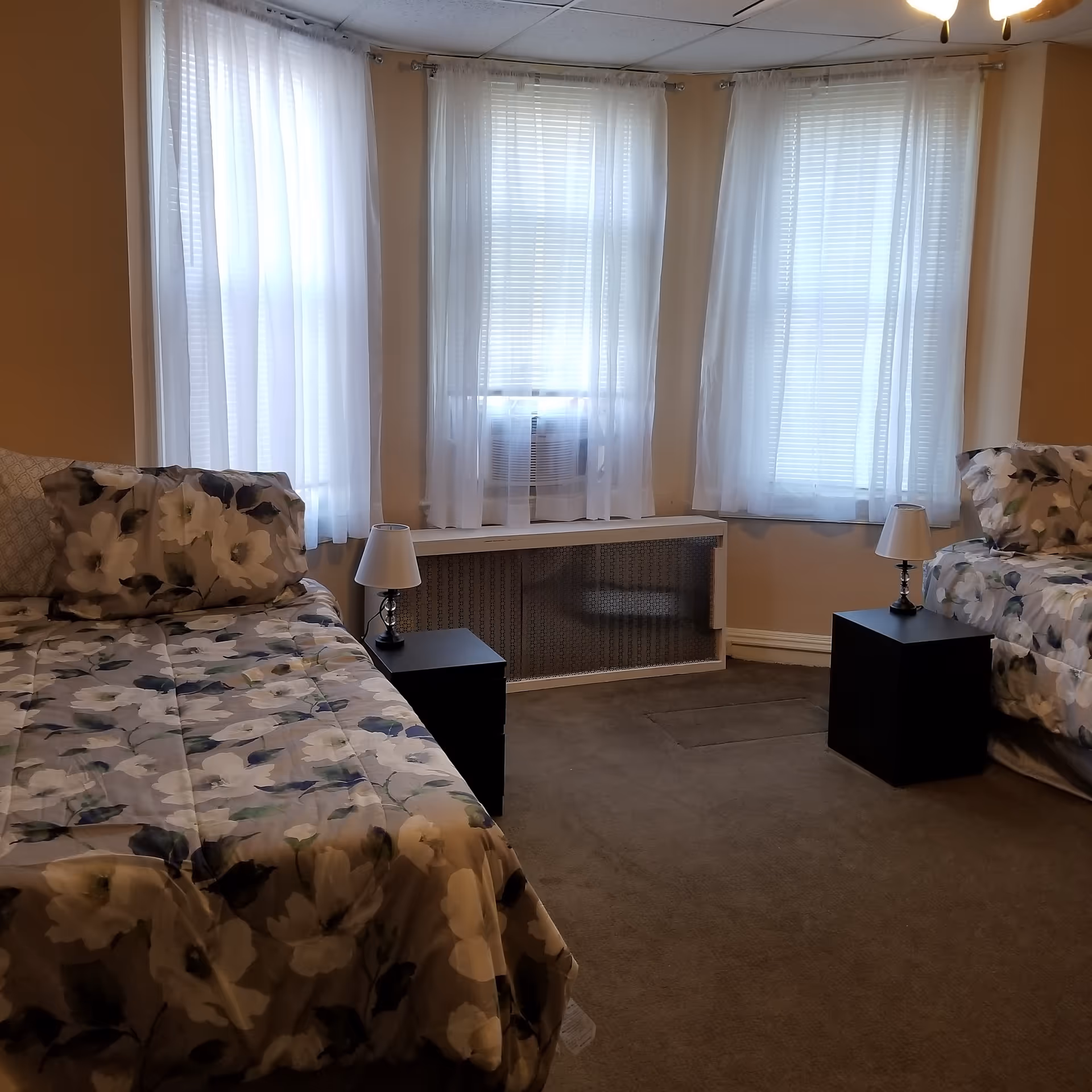 Two beds with floral bedding and nightstands by sheer-curtained windows in a bedroom.