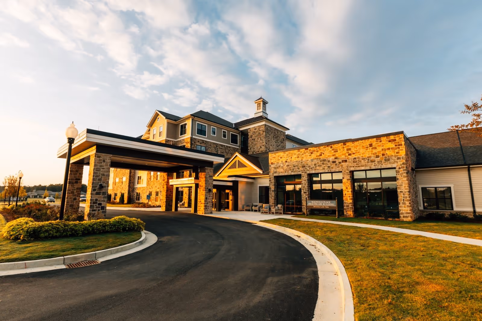 Front entrance of a senior living facility with a covered porte-cochère, stone facade, curved driveway, and landscaped lawn under a partly cloudy sky.