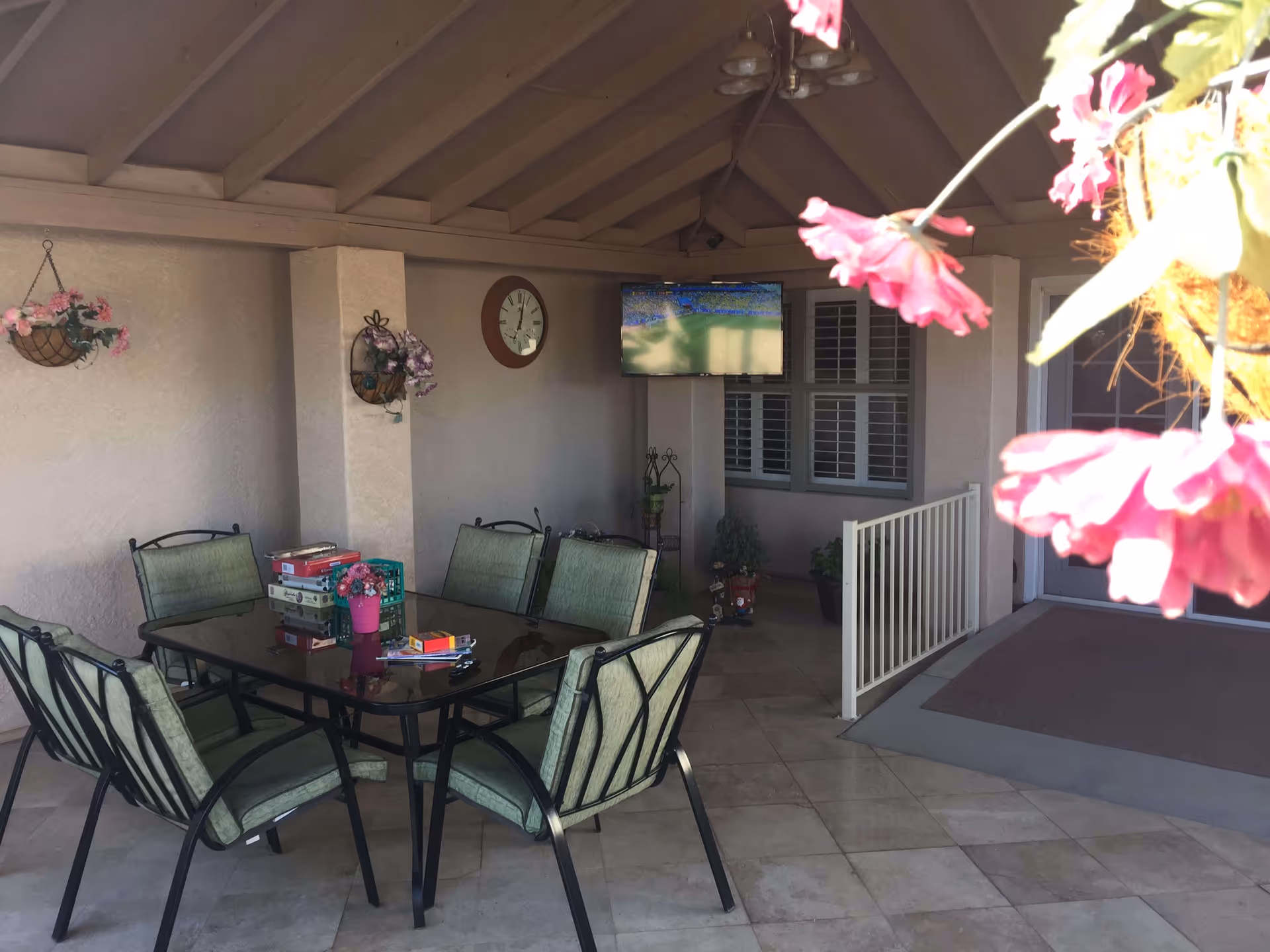 Covered outdoor patio area with a glass-top table surrounded by six green cushioned chairs. On the table are several board games and a small pink flower pot. The patio has a tiled floor, hanging flower baskets, a wall clock, a mounted TV, and a railing leading to a door with windows.