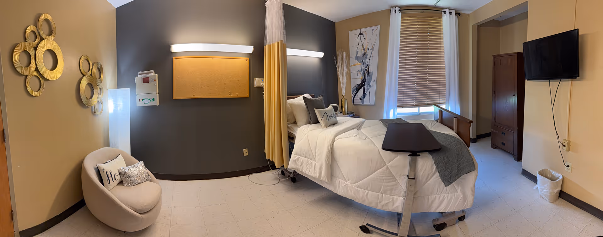 Medical-style bedroom with adjustable hospital bed, window blinds, dresser, TV, and cozy chair.