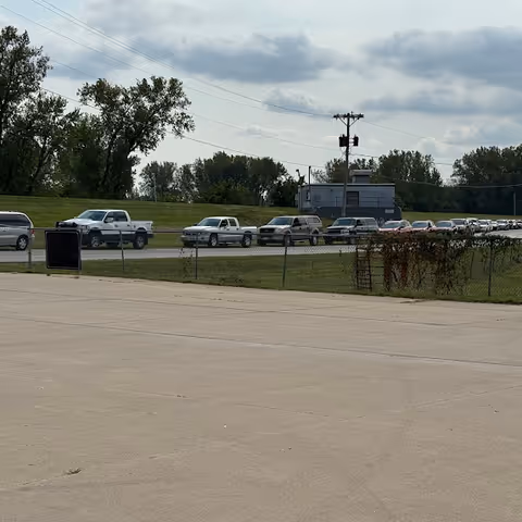 A parking lot scene with multiple cars behind a chain-link fence and utility structures.