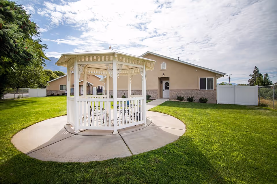 White gazebo on a circular concrete pad in a grassy yard in front of a single-story beige building.