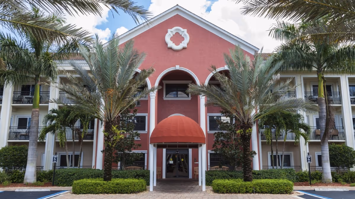 Front exterior view of a three-story building with a red and white facade, palm trees, and a covered entrance with a red awning at Tuscany Villa of Naples.