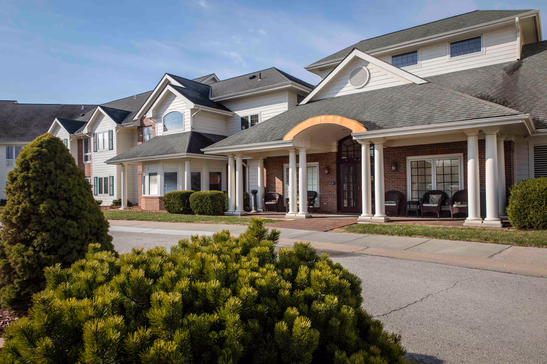 Exterior view of a two-story assisted living facility with a covered entrance supported by white columns. The building has a combination of brick and light-colored siding, with several windows and a paved driveway. Green shrubs and trees are visible in the foreground under a clear sky.