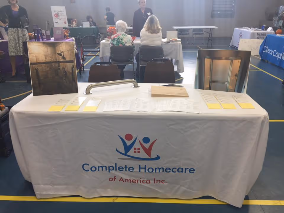 Exhibition table with Complete Homecare of America Inc. logo at a community event indoors.