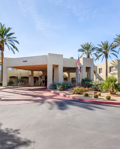 Exterior view of a senior living facility entrance with a covered drop-off area, surrounded by palm trees and desert landscaping including cacti and flowering plants under a clear blue sky.