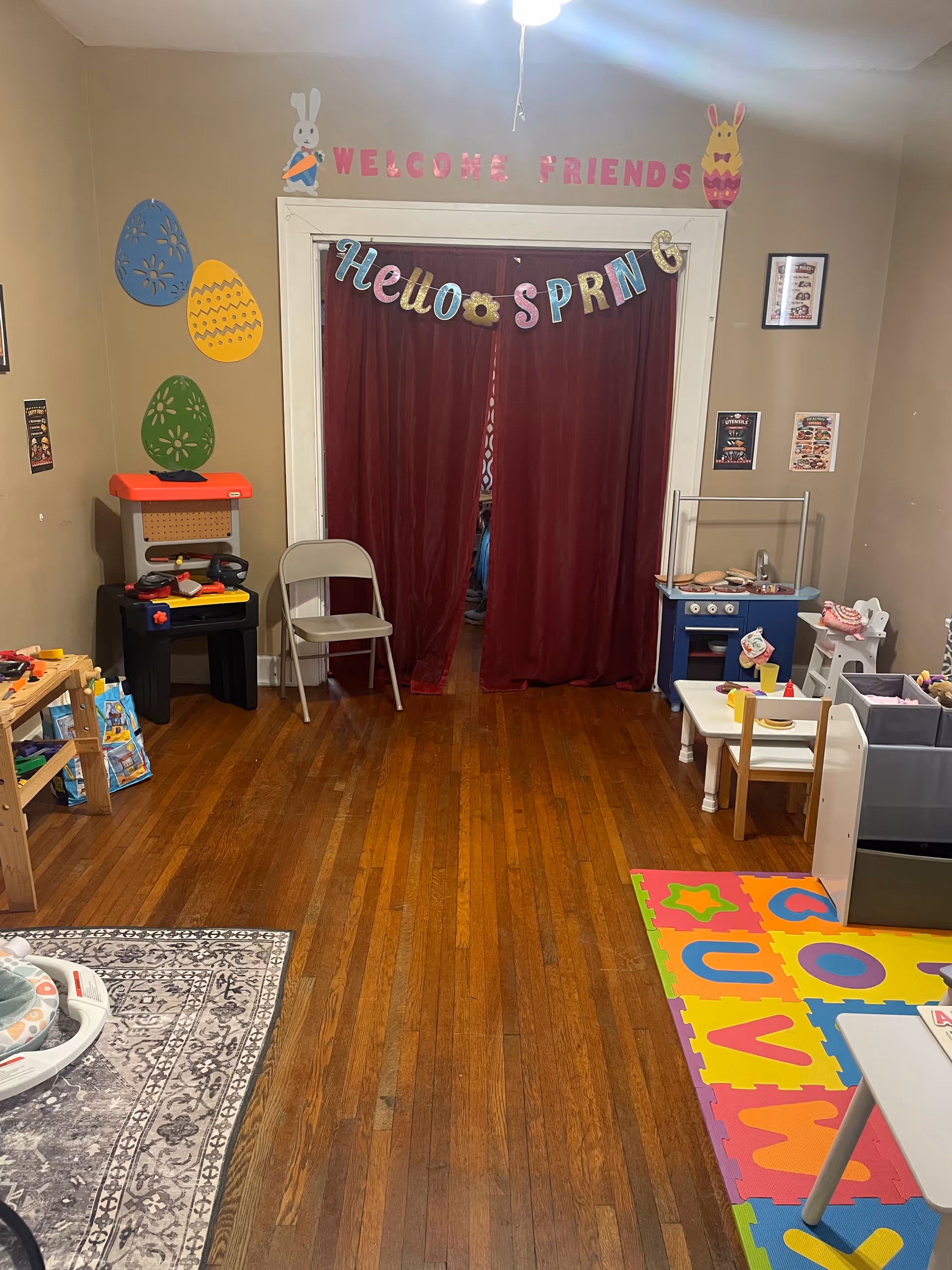 Colorful indoor playroom with toys, a kitchen set, bright mats, and Welcome Friends banner.