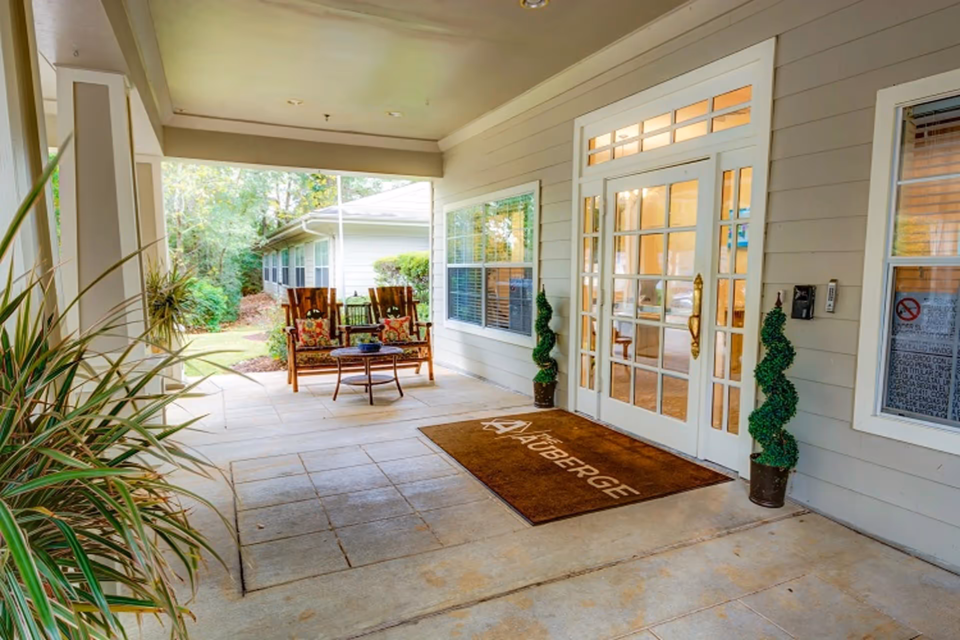 Covered front entrance patio with seating, potted topiaries, and glass double doors with a doormat labeled 'Auberge'.