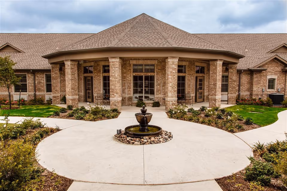 Brick senior living facility entrance with a circular driveway and fountain in front.