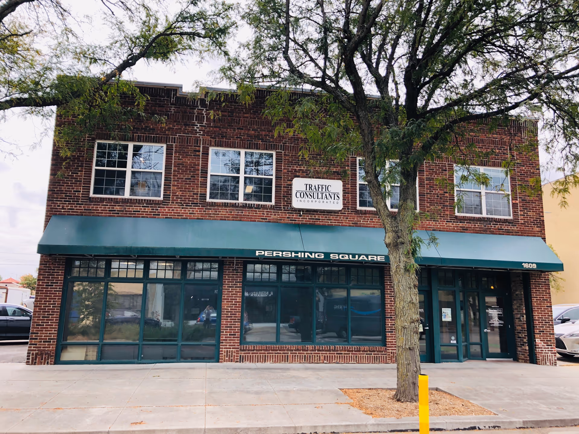 Brick two-story building with green awning, large storefront windows, and sidewalk in front.