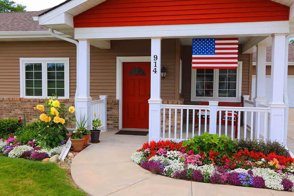 Front entrance of a house-like building with a red door, white porch railing, an American flag, and colorful flower beds.