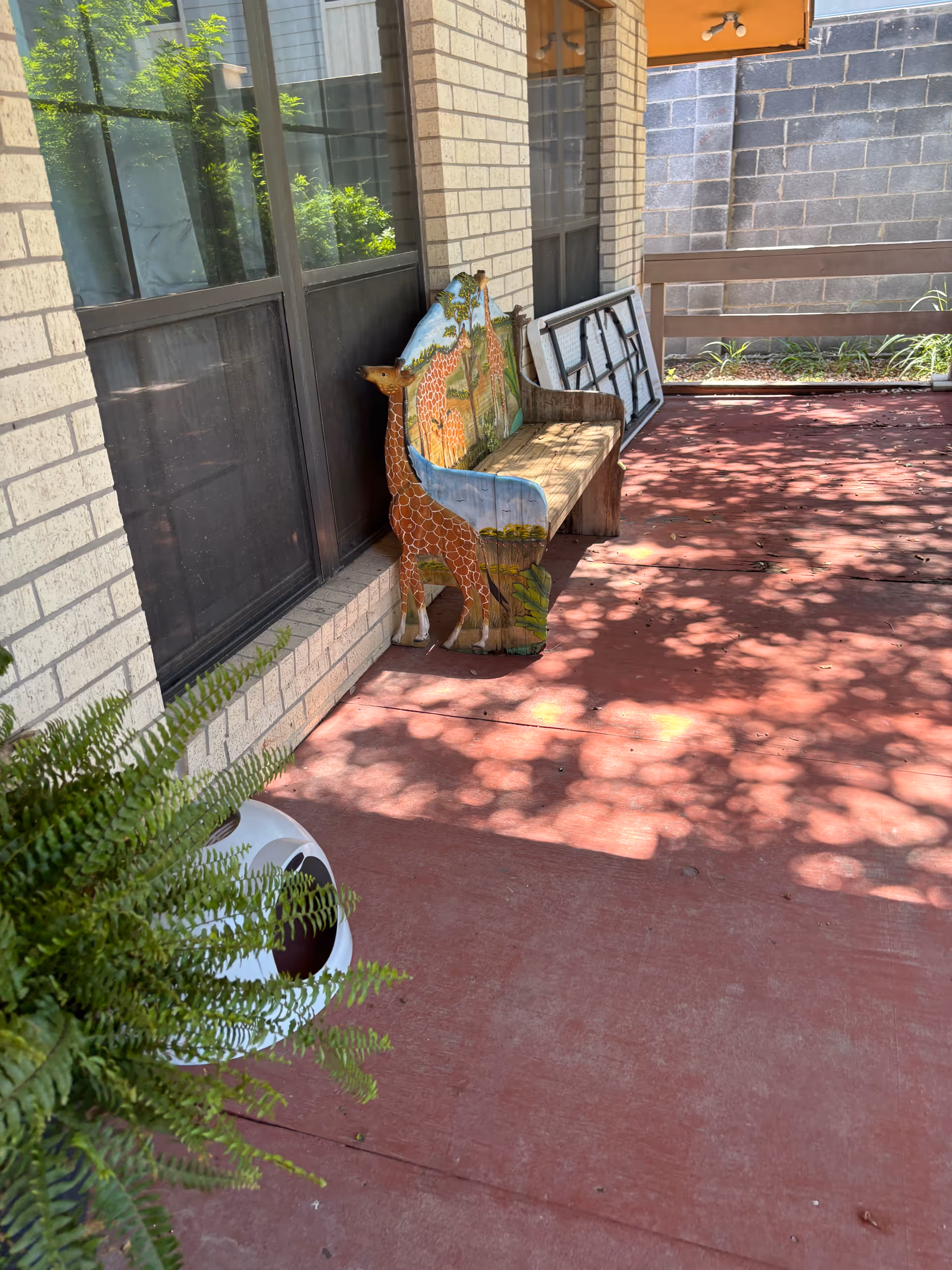 Outdoor patio at a senior facility with a giraffe-themed painted chair, bench, railing, and fern.