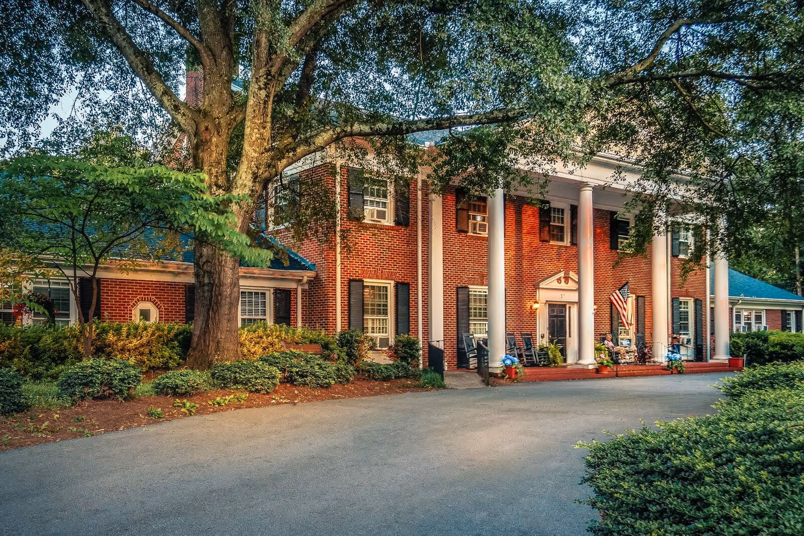 Front view of a red brick building with white columns, a porch with rocking chairs and an American flag, surrounded by trees and landscaping.