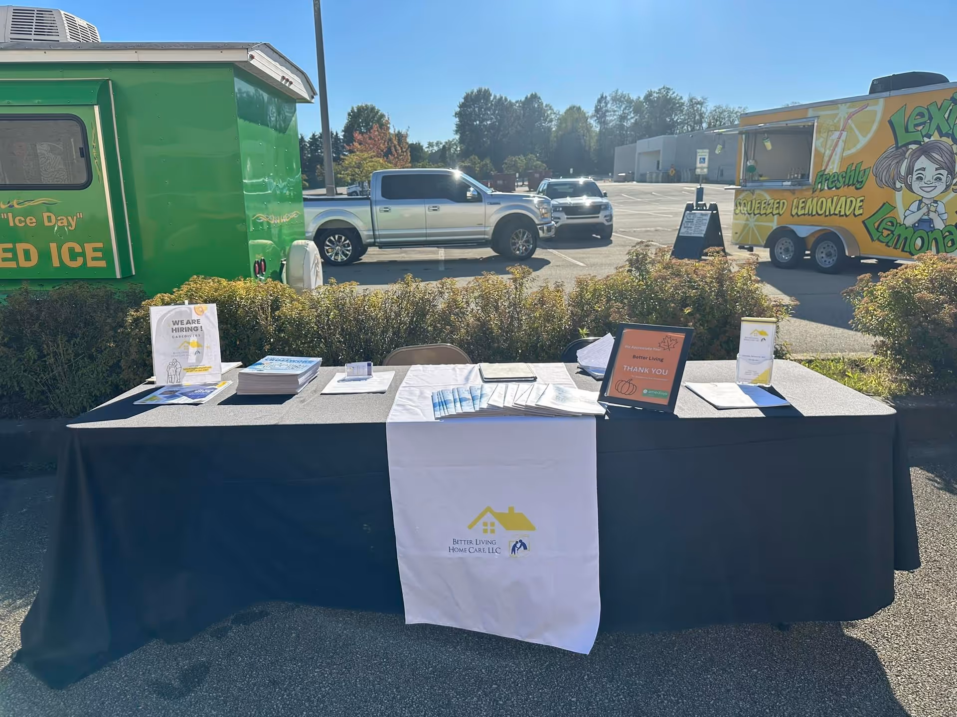 Outdoor recruitment table with brochures and signage for Better Living Home Care in a parking lot.