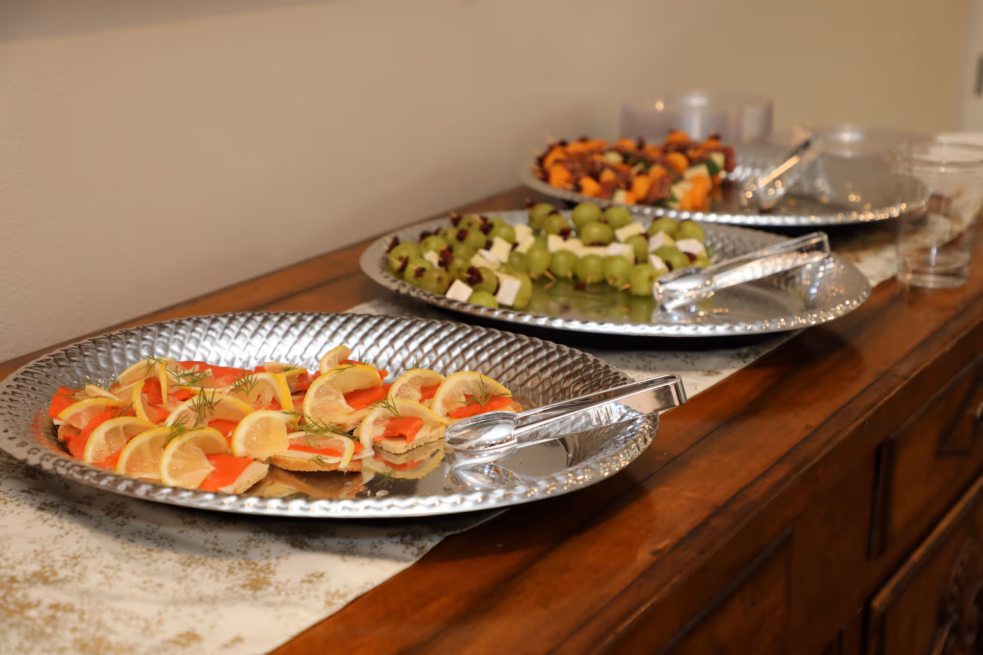 Three silver trays on a wooden sideboard with assorted appetizers including lemon and smoked salmon on crackers, grape and cheese skewers, and fruit and meat skewers, with clear plastic cups and serving tongs nearby.