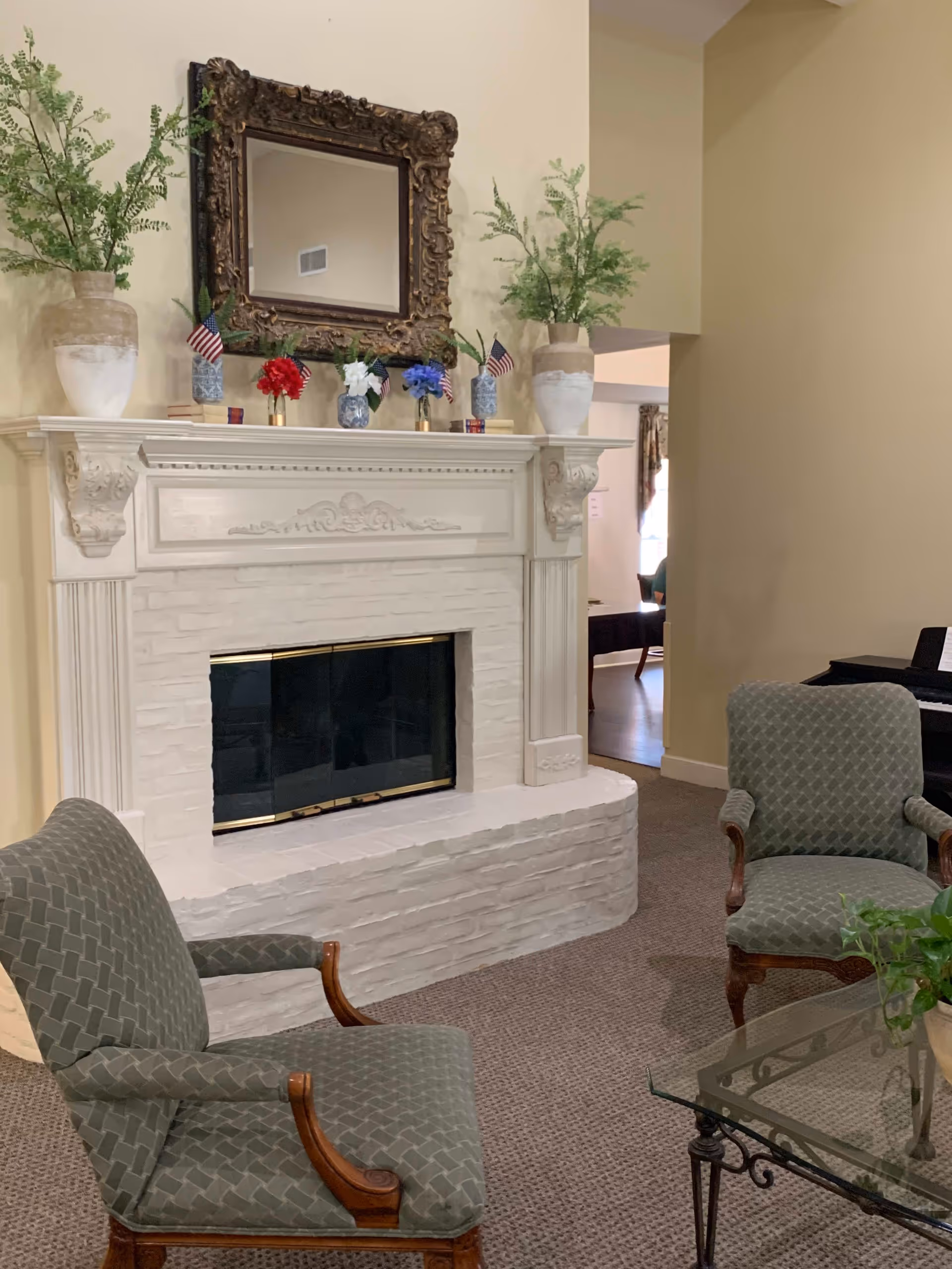 A cozy living room area featuring a white brick fireplace with an ornate dark-framed mirror above it. The mantel is decorated with two large vases with greenery, small American flags, and red, white, and blue floral arrangements in small vases. Two upholstered armchairs with wooden armrests are positioned facing each other near a glass coffee table with a potted plant. The room has beige walls and carpeted flooring.