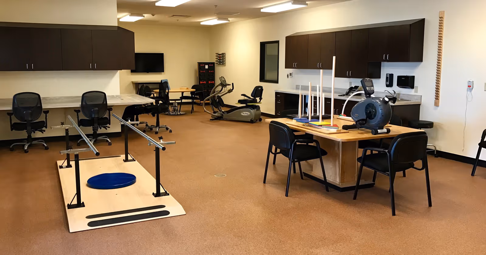 A rehabilitation or physical therapy room with exercise equipment including a stationary bike, parallel bars for walking practice, therapy tables with chairs, and cabinets along the walls. The room has a clean, organized appearance with brown flooring and white walls.