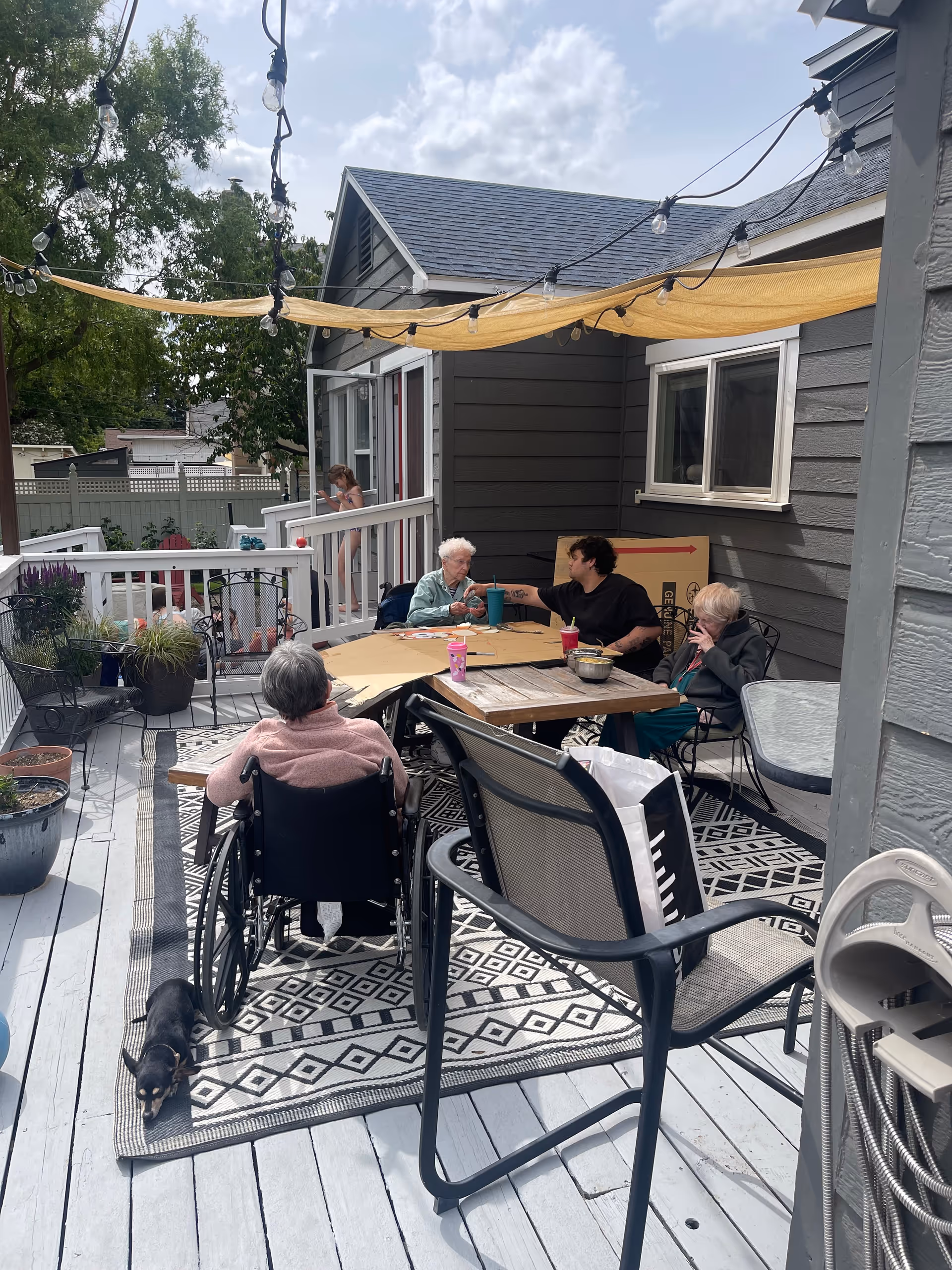 Senior residents socialize around a patio table on a wooden deck outdoors; a cat rests nearby.