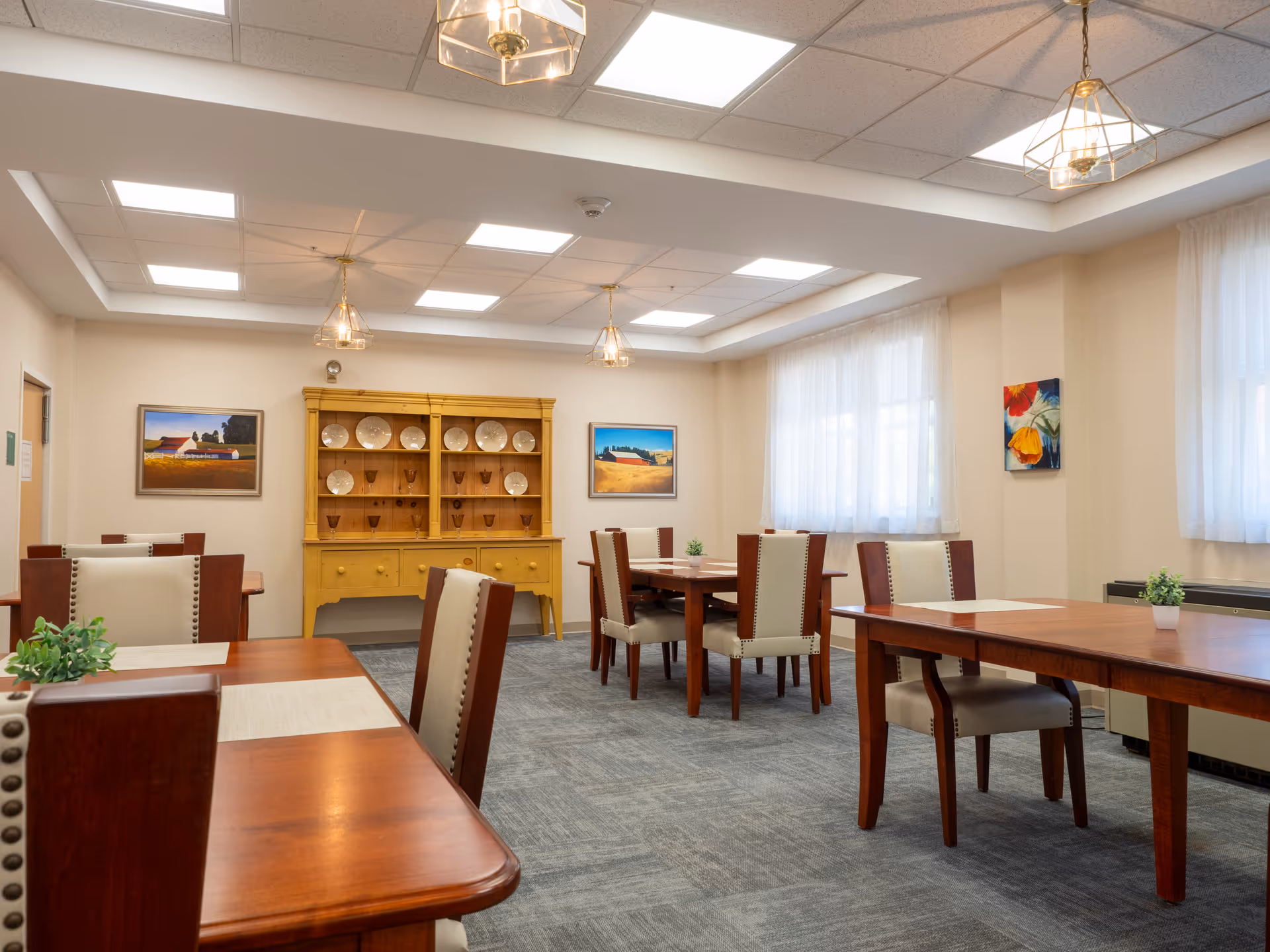 Well-lit senior living dining room with wooden tables, chairs, and a china cabinet.