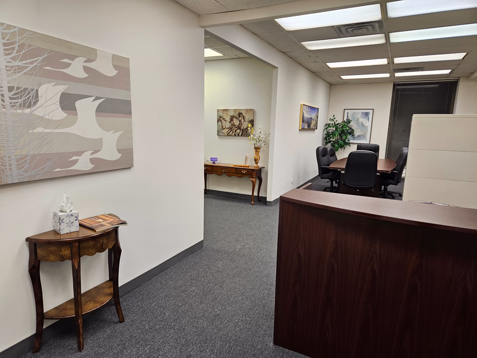 Reception desk in a hallway with wall art, side table, and a conference room visible.