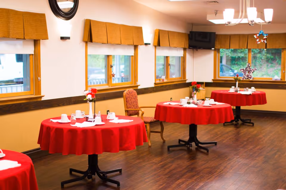 Dining room with round tables covered in red tablecloths, set with white cups, glasses, and napkins. The room has wooden floors, multiple windows with brown valances, a wall-mounted TV, and a chandelier with white lampshades.