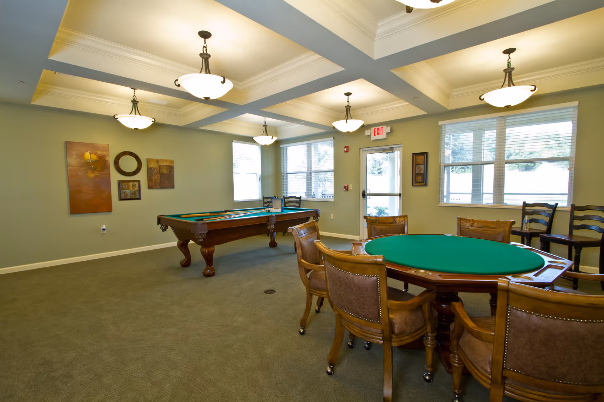 A well-lit game room with a pool table and a round card table surrounded by chairs. The room has green carpet, light green walls, decorative ceiling lights, and several windows letting in natural light. There are framed artworks on the walls and an exit door with a red exit sign above it.