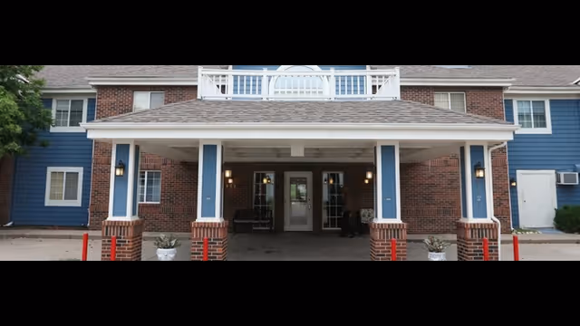Front exterior view of a two-story building with a covered entrance supported by brick and blue pillars. The building has a combination of red brick and blue siding with multiple windows and a white balcony above the entrance.