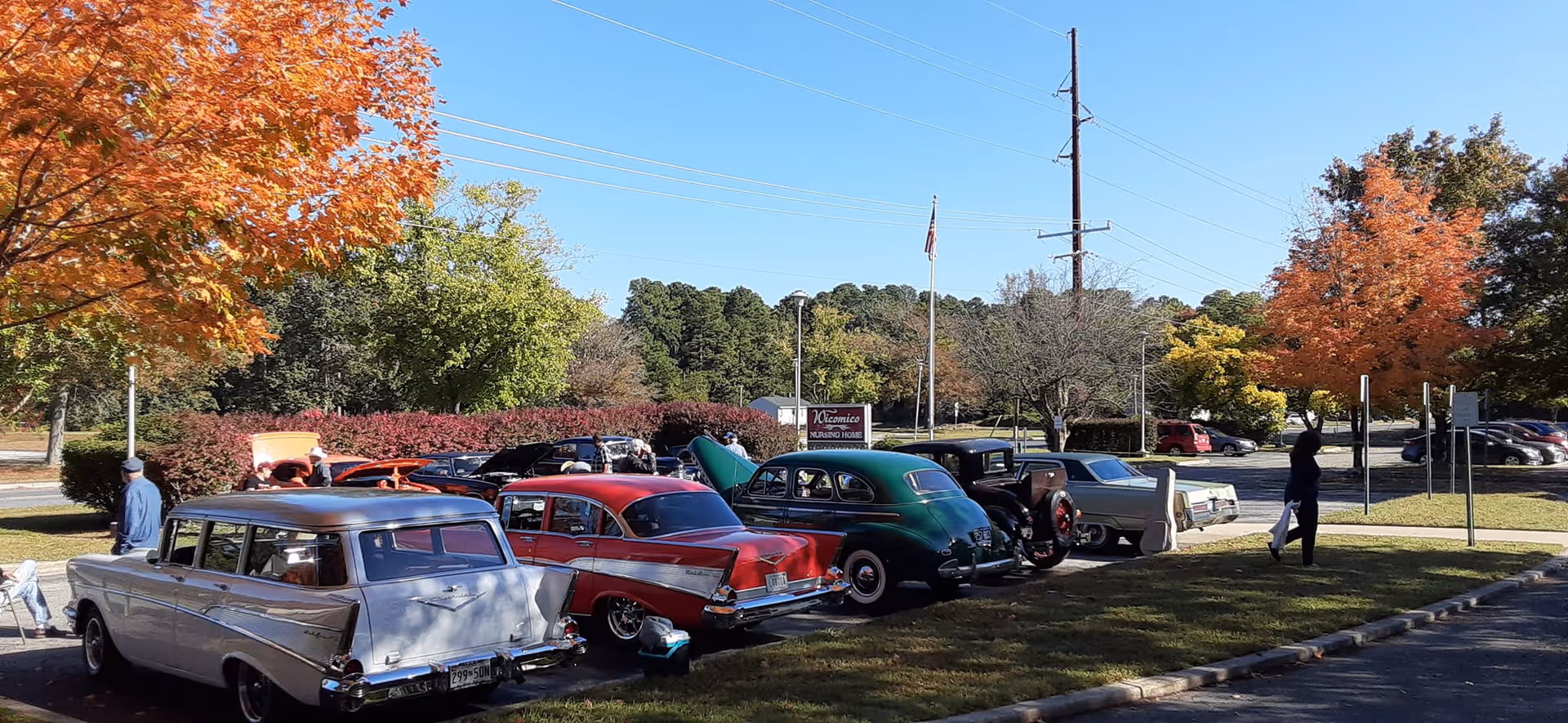 Rows of vintage cars parked outside a nursing home beneath autumn trees and clear sky