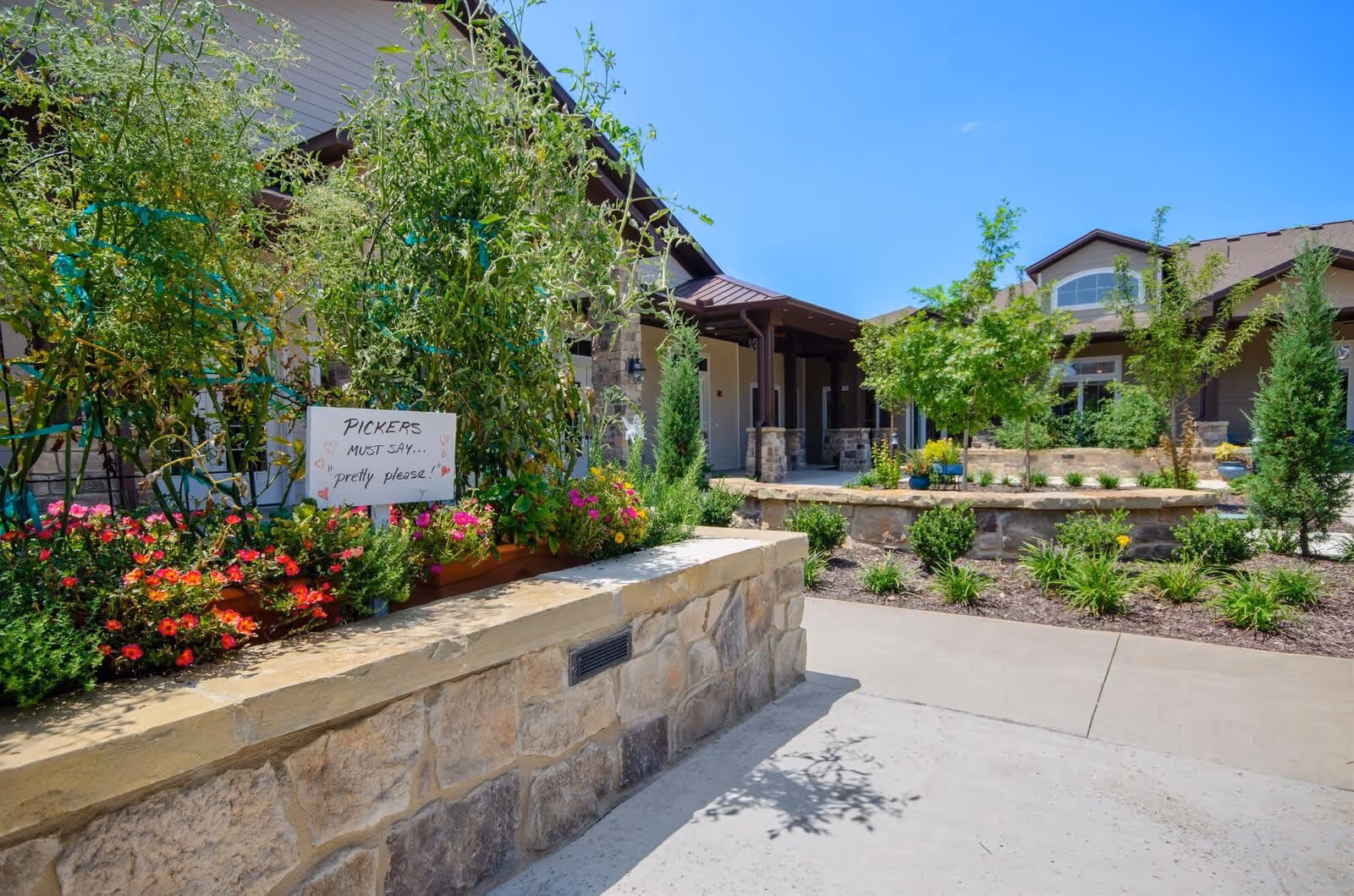 Outdoor garden area at Grand Brook Memory Care of Fishers featuring raised stone flower beds with colorful flowers and green plants, a sign that reads 'PICKERS MUST SAY... pretty please!', and a building with a covered porch in the background under a clear blue sky.