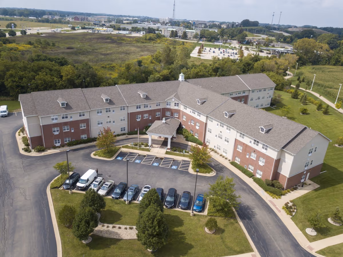 Aerial view of a three-story senior living building with a covered entry, circular driveway and parked cars surrounded by lawns and trees.