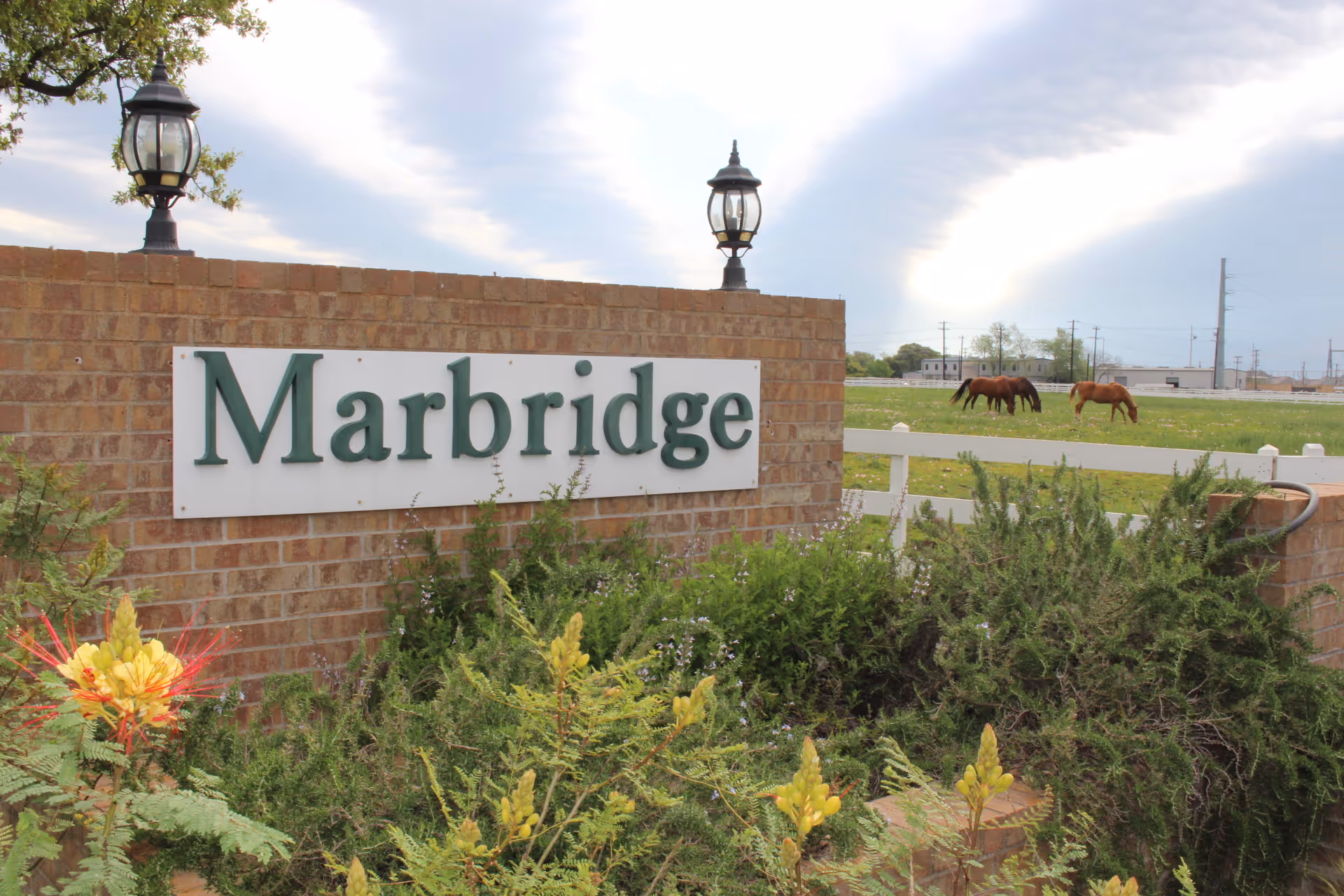 Bricked entrance with Marbridge sign, lamps, greenery, and distant horses.