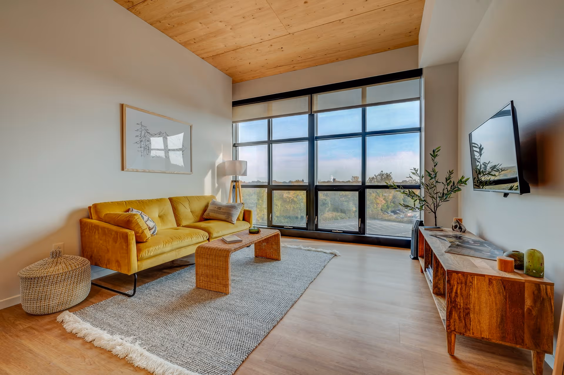 A modern living room with a mustard yellow sofa, a woven coffee table on a gray rug, a floor lamp, and a wooden media console with decorative items. Large windows provide a view of trees and a parking lot outside.